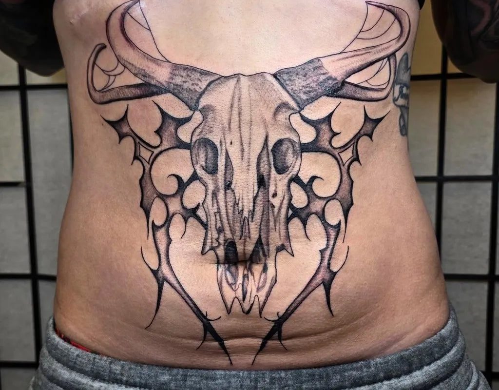 여성 복부에 새겨진 커다란 블랙워크 롱혼 두개골과 부족 문양 날개 문신