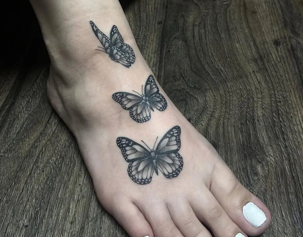 여성 발에 새겨진 작고 검은색과 회색의 나비 세 마리 문신